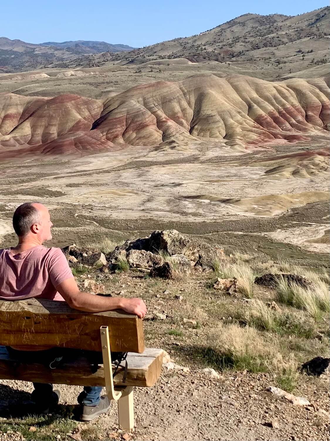 Matthew Kessi sits on a bench overlooking the beautiful rolling pattern of the Painted Hills in Central Oregon. The colors of red and yellow flow in gentle lines below a blue sky. 