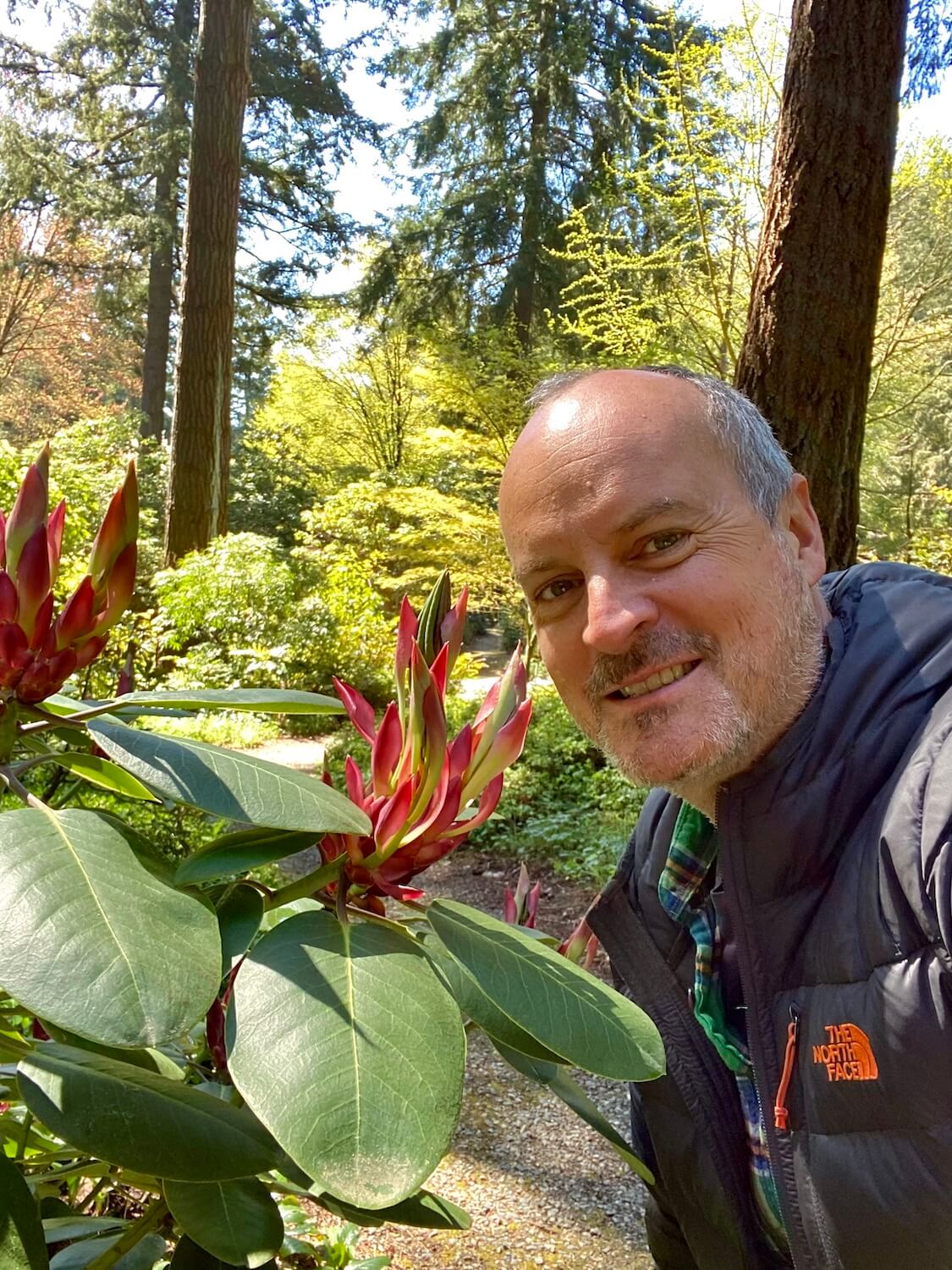 Matthew Kessi poses for a selfie next to a nearly budding Rhododendron plant, not yet fully formed.  He is smiling and wearing a gray down jacket. 