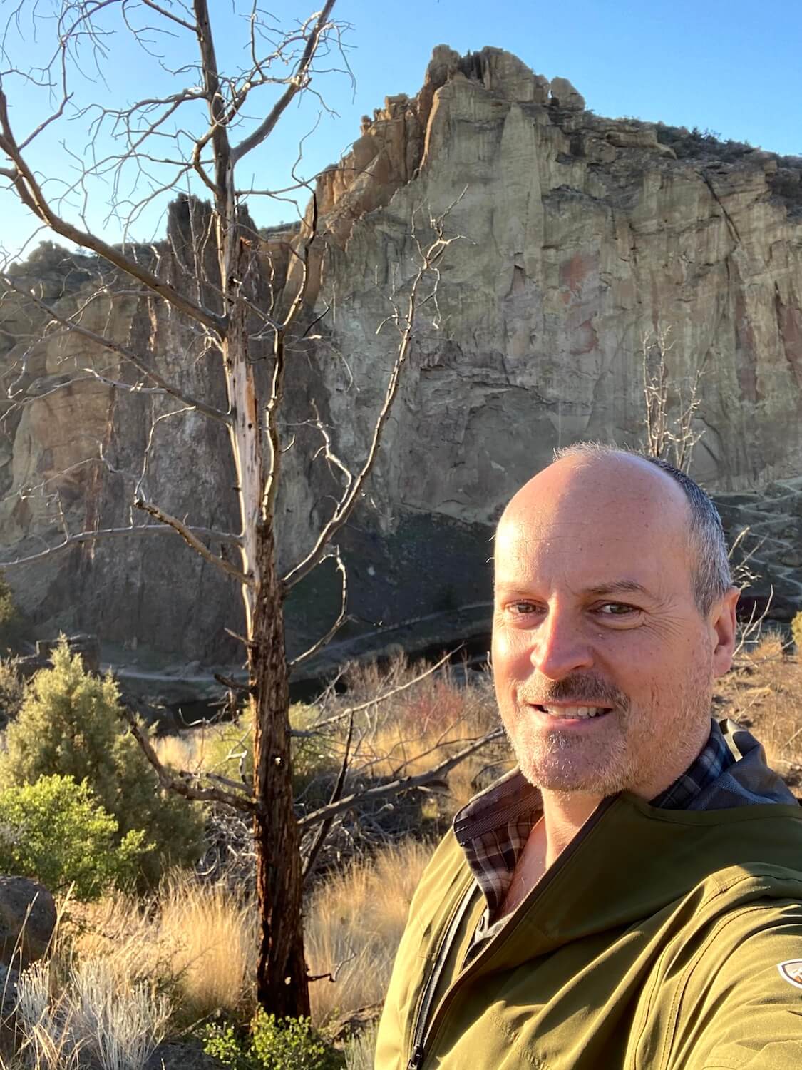 Matthew Kessi takes a selfie of himself standing in front of a rising rock face at Smith Rock State Park near Redmond, Oregon.  He's wearing a green coat and plaid shirt and stands in front of a dead juniper tree that stands amongst low brush and grass. 