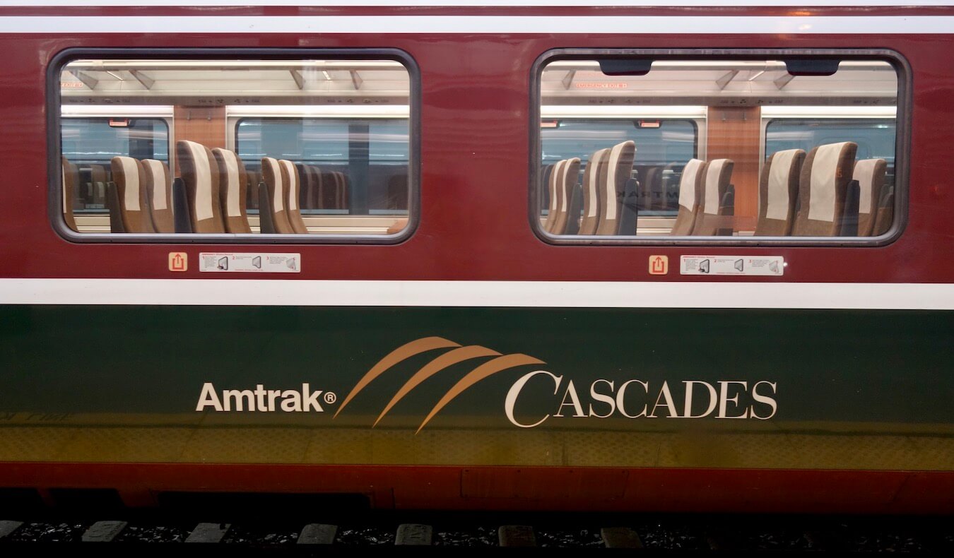 The exterior of an Amtrak Cascades train from Seattle to Vancouver shows the logo of the train on top of a green stripe with brown coloring around the windows.  
