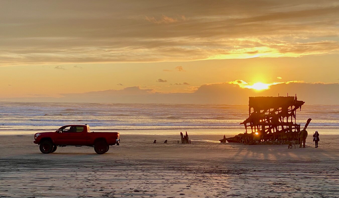 The sun sets in the background shining golden hues on the shipwreck of the Peter Irdale, near Astoria Oregon.  A red truck drives along the beach as visitors view the twisted metal of the shipwreck.  