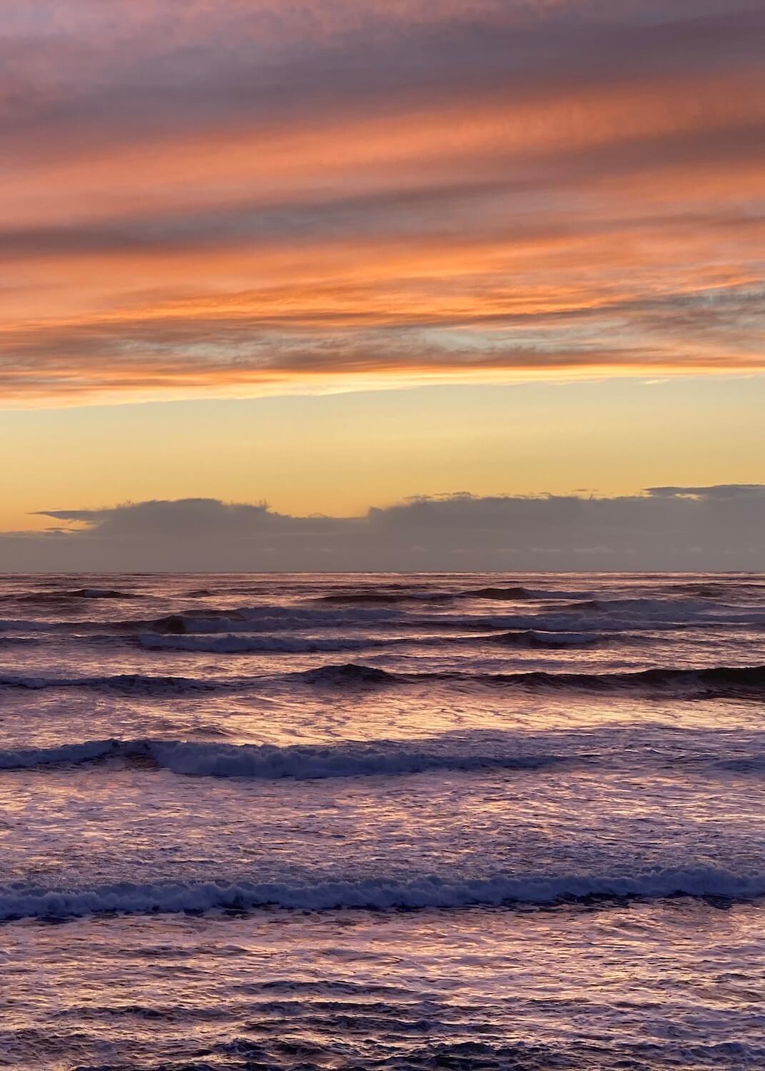 The sunsets on the Pacific Ocean creating beautiful shades of purple and pink amongst the foaming layers of waves pushing toward the sandy beach. 