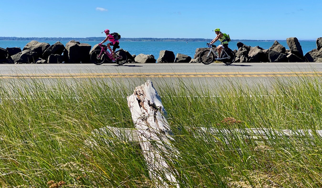 Two bicyclists ride along a coastal road with large boulders used as a barricade from the water in the distance. 