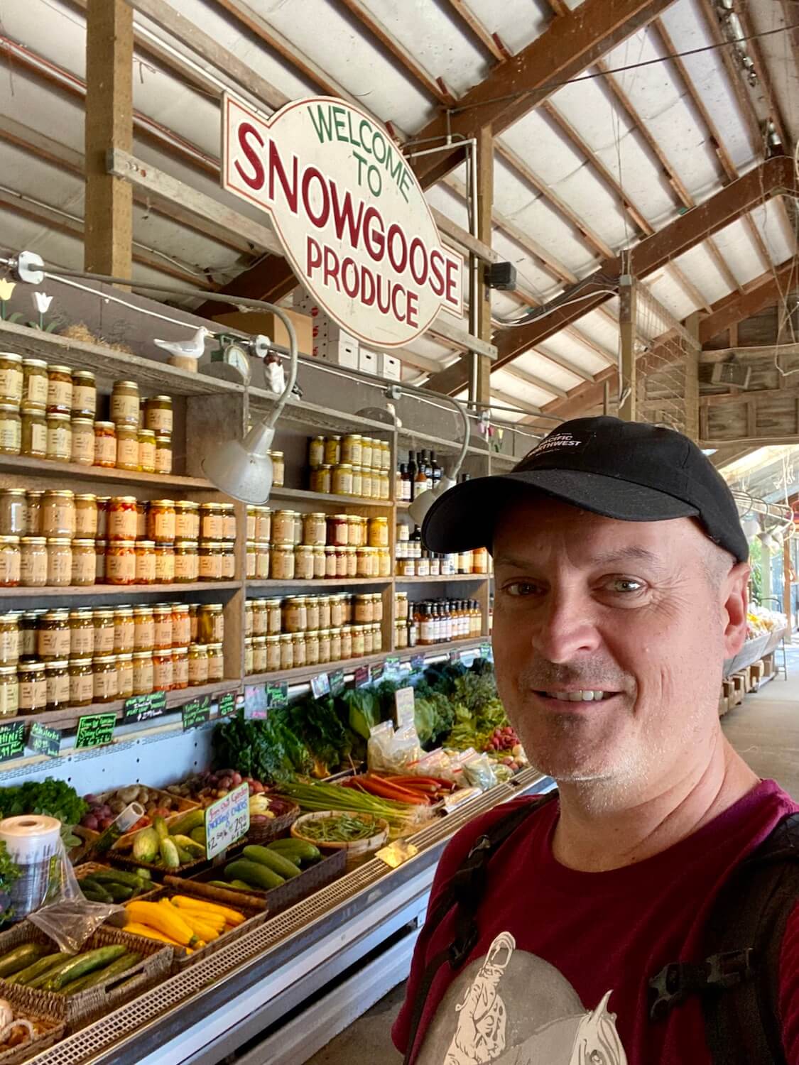 Matthew Kessi poses for this selfie at Snow Goose Produce in the Skagit Valley. He's wearing a magenta t-shirt and black cap and there are shelves of farm products on the wall behind him, as well as vegetables in a cooler below.