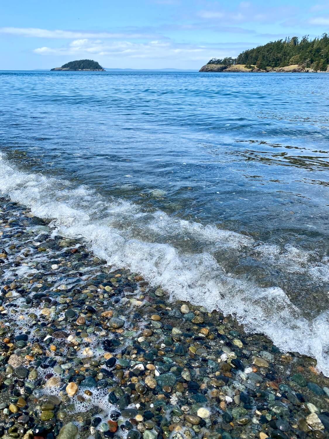 The waves of the Salish Sea at Deception Pass State Park crash down on the pebbles on the beach while rocky fir-tree covered islands are seen in the background. 