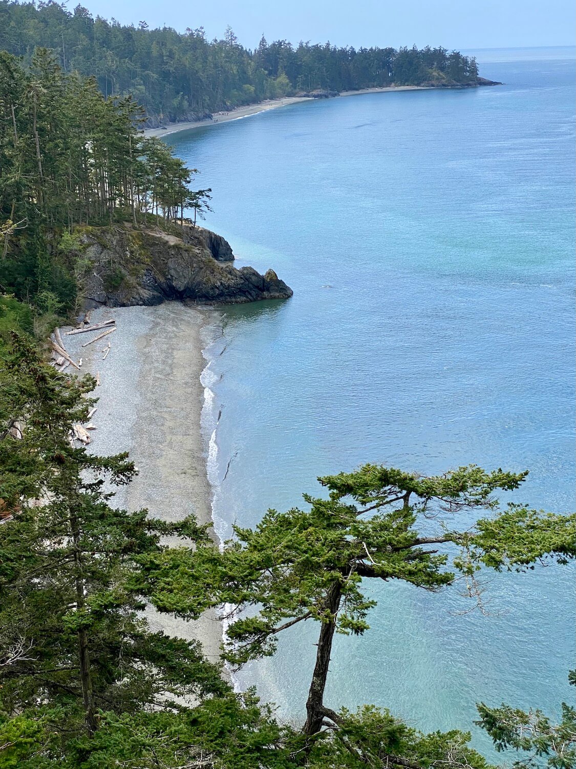 A dramatic view of North Beach at Deception Pass State Park shows long spans of pebble beaches with rocky cliffs, covered with scraggly fir trees jutting out into the sea.  