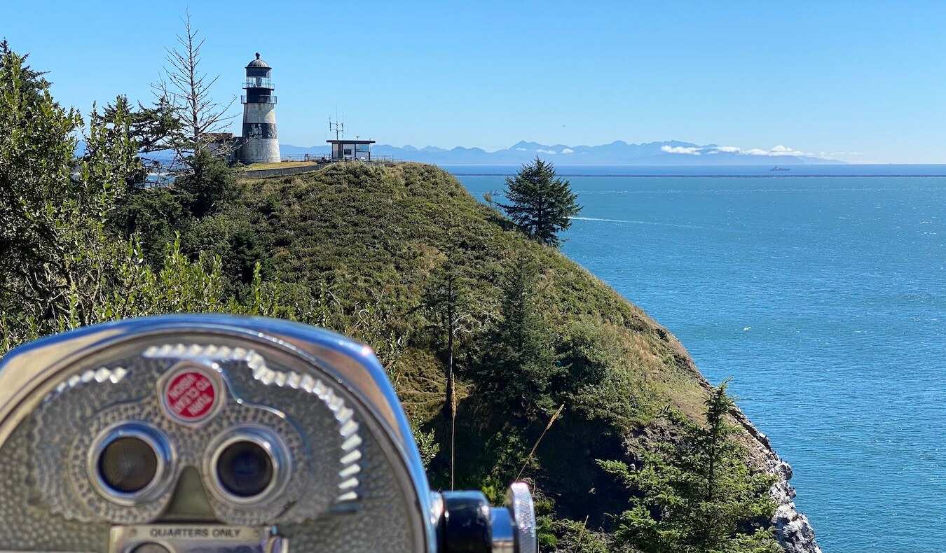 A shiny metal viewfinder is focused on a lighthouse on an adjacent hill while rolling mountains rise up across the mouth of the Columbia River at Cape Disappointment. 