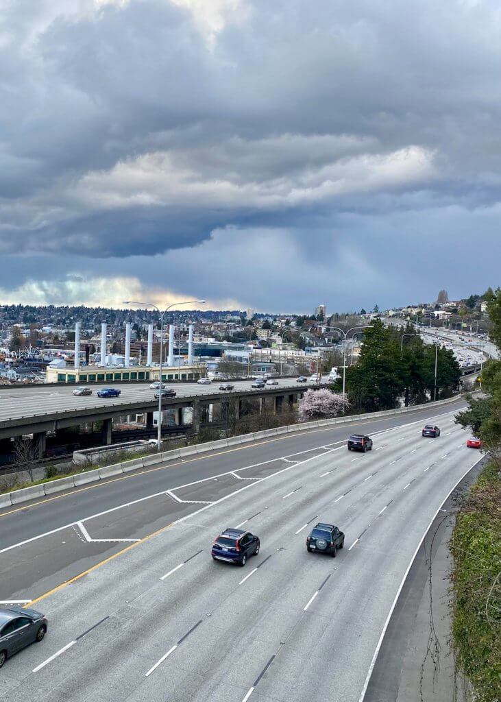 This photo shows Interstate 5 in Seattle with five lanes of traffic but only a few cars. This is the start of the trip from Seattle to Vancouver. In the distance, the cloudy skies are dramatic as if a thunderstorm will start soon.