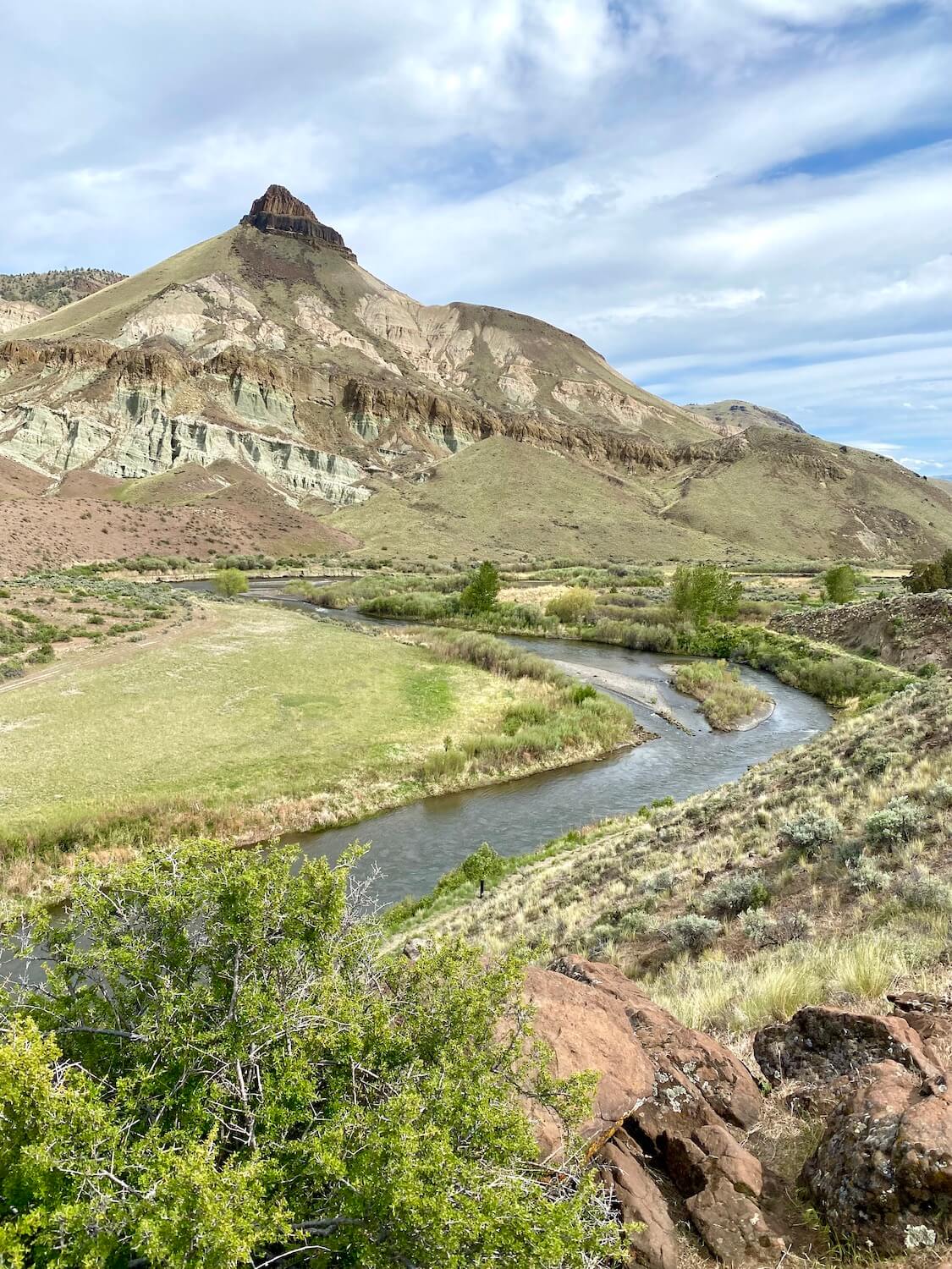 This photo shows the rocky mountain point of Sheep Rock which is in the John Day Fossil Bed National Monument.  The river flows through the photo while green farmland flows from the bank towards the rocky canyon above.