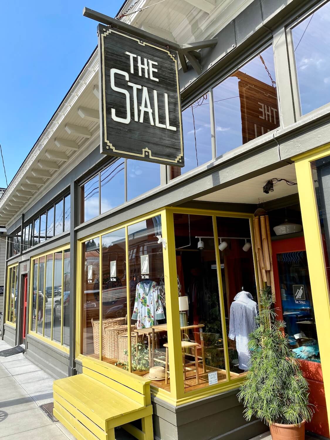 The Stall is a boutique on the Main Street of La Conner, Washington.  The window frames are painted a bright color of yellow and you can see various clothing options inside the window. There is also a yellow bench on the sidewalk leaned up against the windows of the shop.