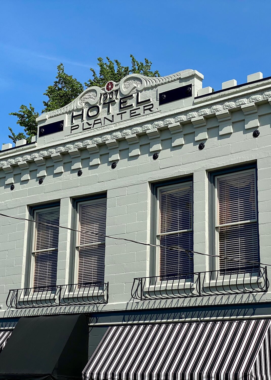 Hotel Planter, in downtown La Conner, Washington is a historic hotel that was built in 1907.  The facade is an olive green with black trim around the windows.  The sky above is blue and there is a green leafy tree above the cornice of the hotel. Covering a window on the first floor is an awning with black and white stripes. 