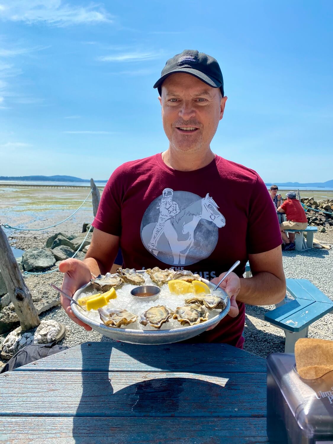 Matthew Kessi poses for a selfie at Taylor Shellfish along the Salish Sea. He's holding a tray of a dozen oysters packed in ice with wedges of Lemon and ramekins of sauce. The sky is sunny and blue in the background.