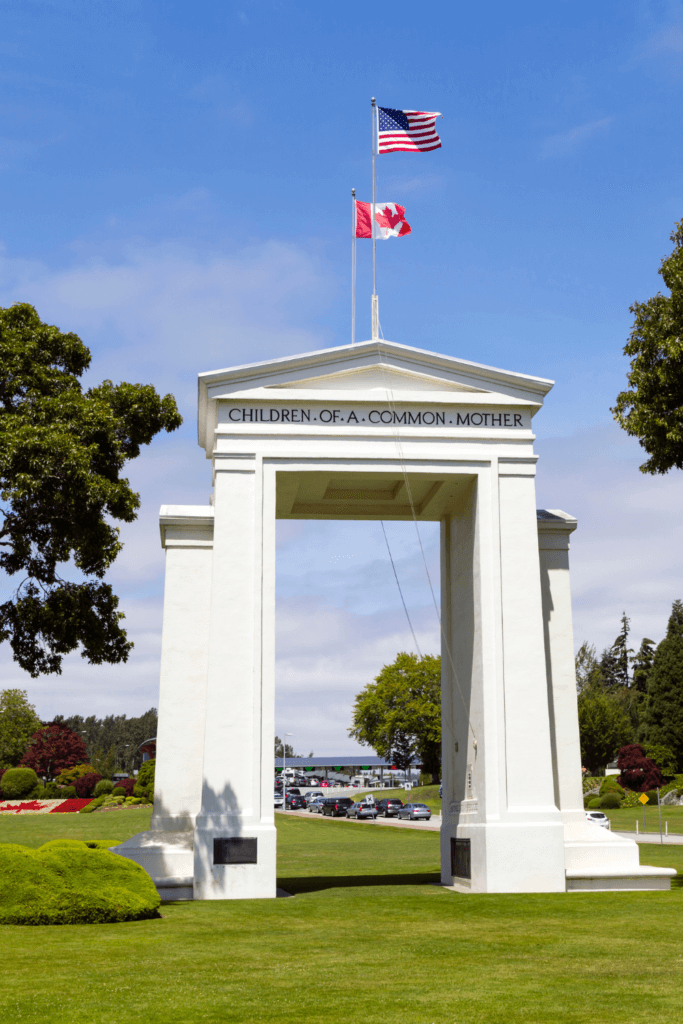 The giant Peace Arch at the United States Canada border is a milestone along the Seattle to Vancouver Drive. The giant white marble columns are located on a grassy green lawn with trees and gardens and both the US and Canadian flags fly proudly overhead.