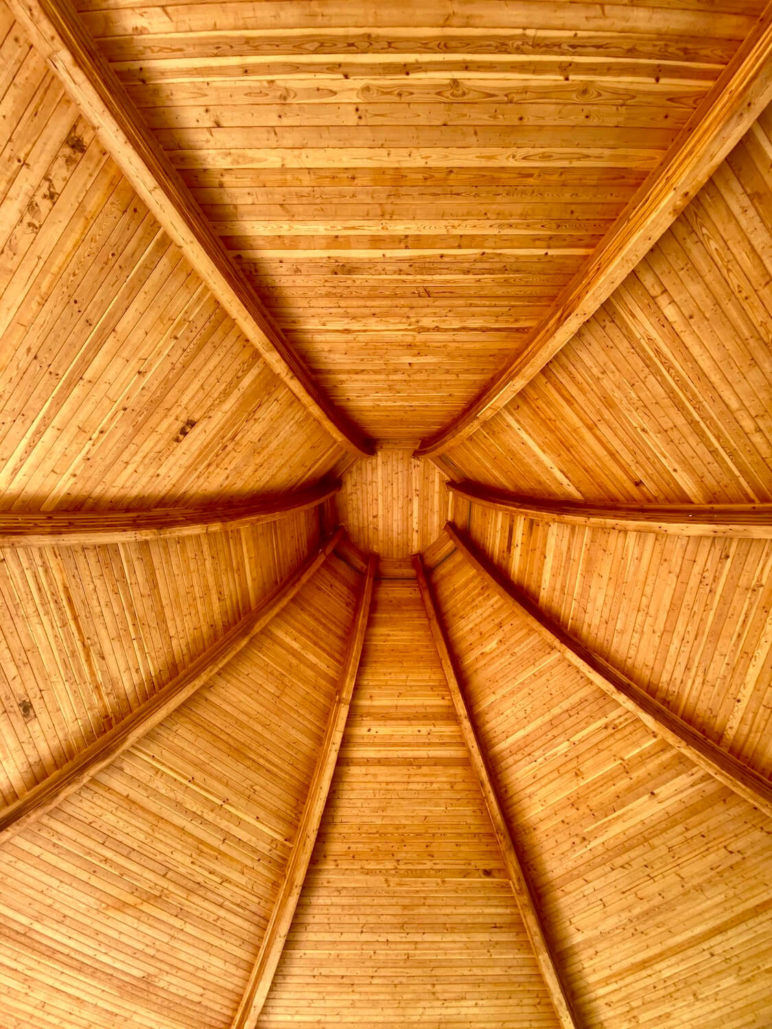 This inside of a pavilion made to resemble a traditional cedar hat worn by the Swinomish Tribe.  The cedar planks are an orangish glow and bend up to the ceiling, which has an octagon shaped cap.  