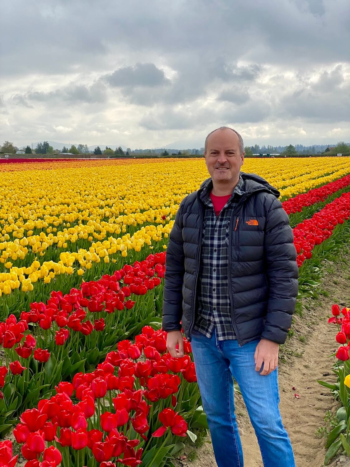 Matthew Kessi stands in front of fields of endless tulips during the Skagit Valley Tulip Festival.  He's wearing a puffy grey North Face jacket and jeans.  