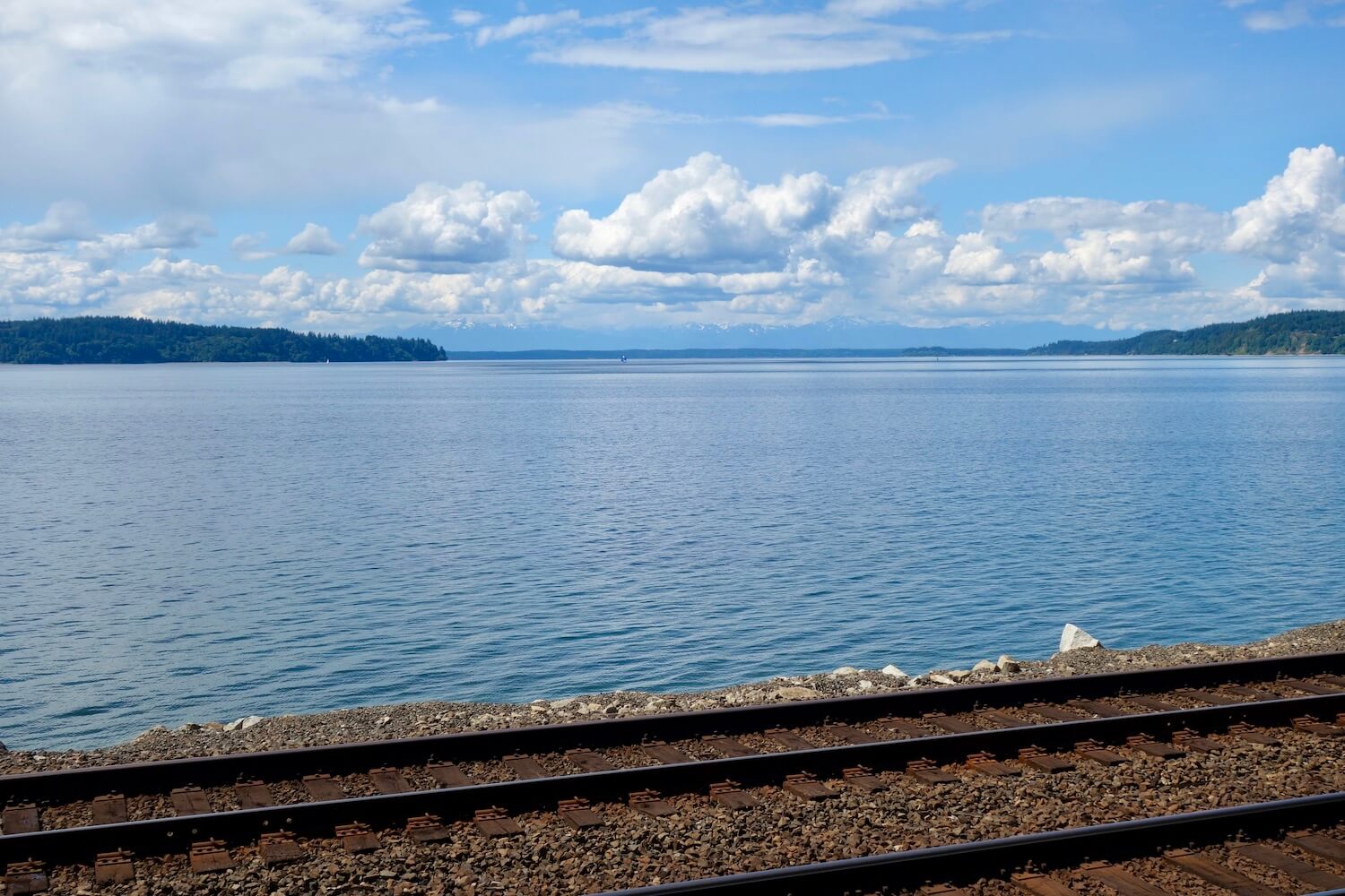 A great shot of the Salish Sea from the Seattle to Portland train.  There are two thick steel rails in a bed of gravel while the blue sea is tranquil beyond with islands and mountains in the far distance. 
