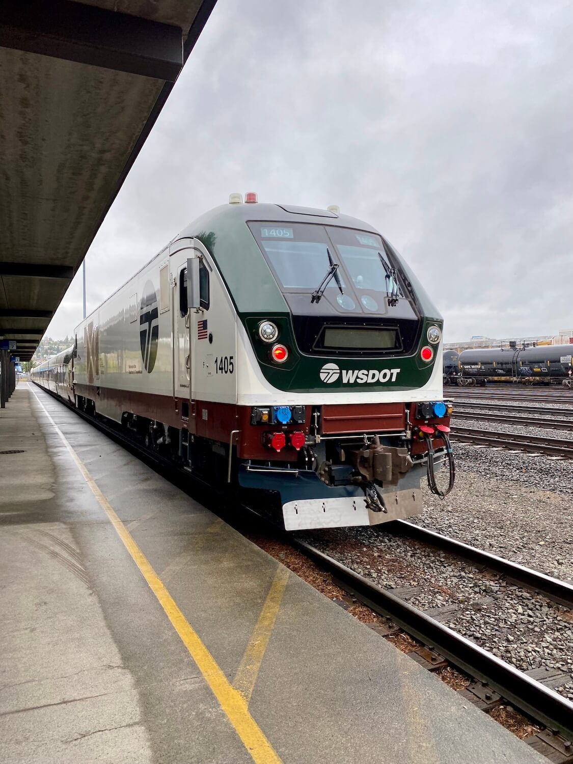 The engine of the Amtrak Cascades on the way from Seattle to Portland. The yellow line of the train platform can be seen running the length of the photo which contacts with the green and white coloring of the train engine that has WSDOT displayed on the front.