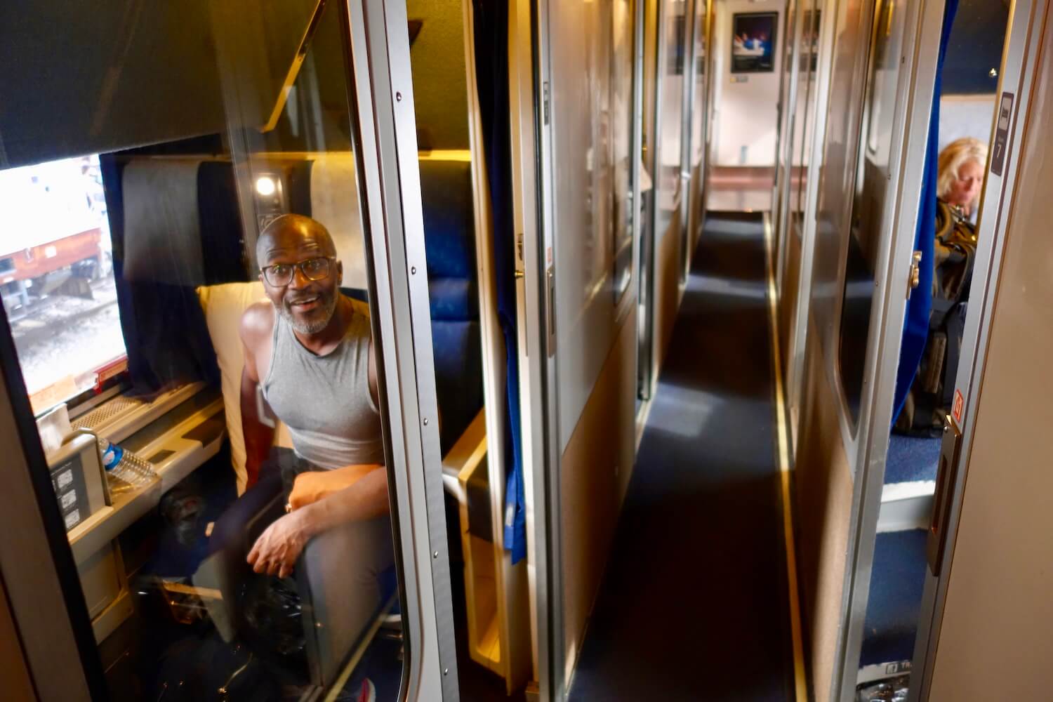 A black man sitting in a roomette compartment on an Amtrak train gets comfortable and looks at the camera, which is in the hallway between the two sides of rooms.  