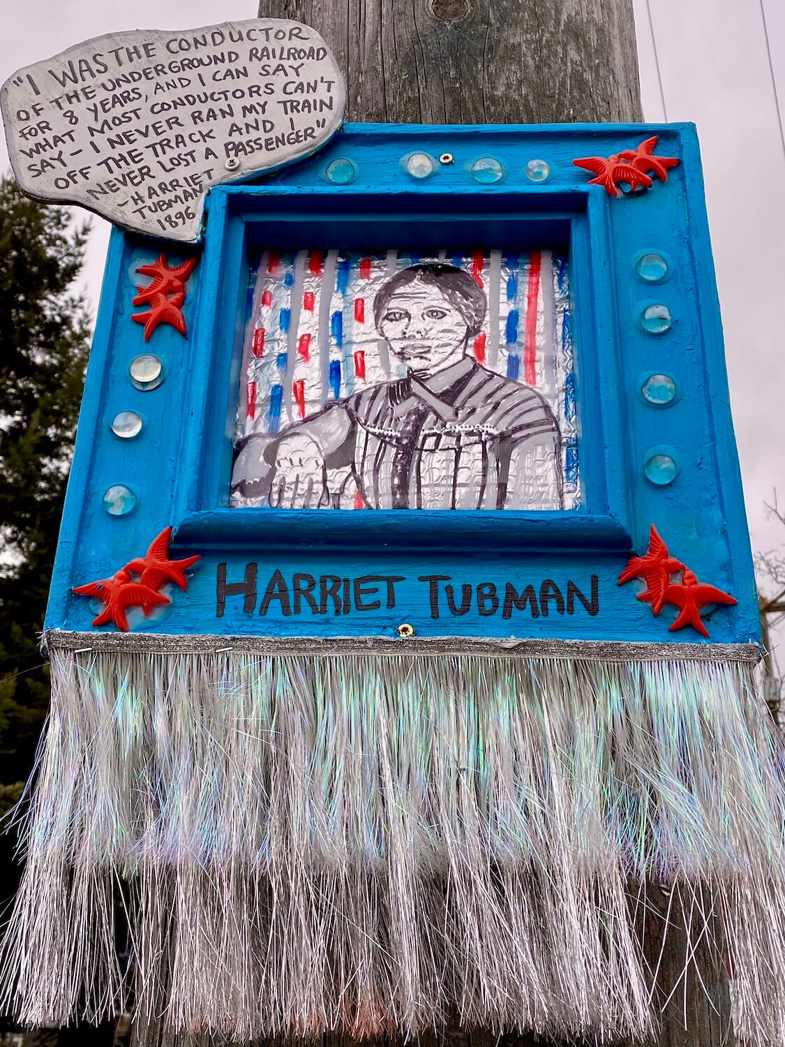 A blue painted frame with glued on birds and clear glass orbs showcases a picture of Harriet Tubman etched into tin foil.  There is a dialog bubble above her explaining her role in equality in America.  There is tinsel hanging from the bottom of this art installment, which was found on a telephone pole in the Columbia City neighborhood of Seattle.  
