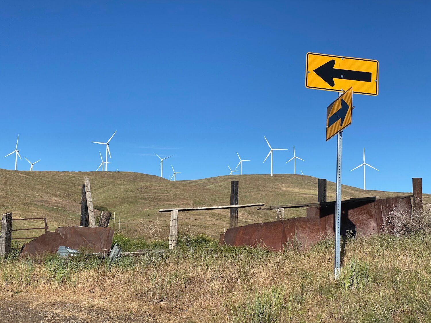 A hillside lined with giant white windmills reaches up to a blue sky while two orange road signs show arrows pointing in opposite directions.  There are pieces of rusted metal leaning up against a barbed wire fence. 