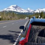 A blue SUV is parked on the side of a highway on an Oregon road trip while the South Sister of the Cascade Mountains rises up snow-covered, toward the blue sky.
