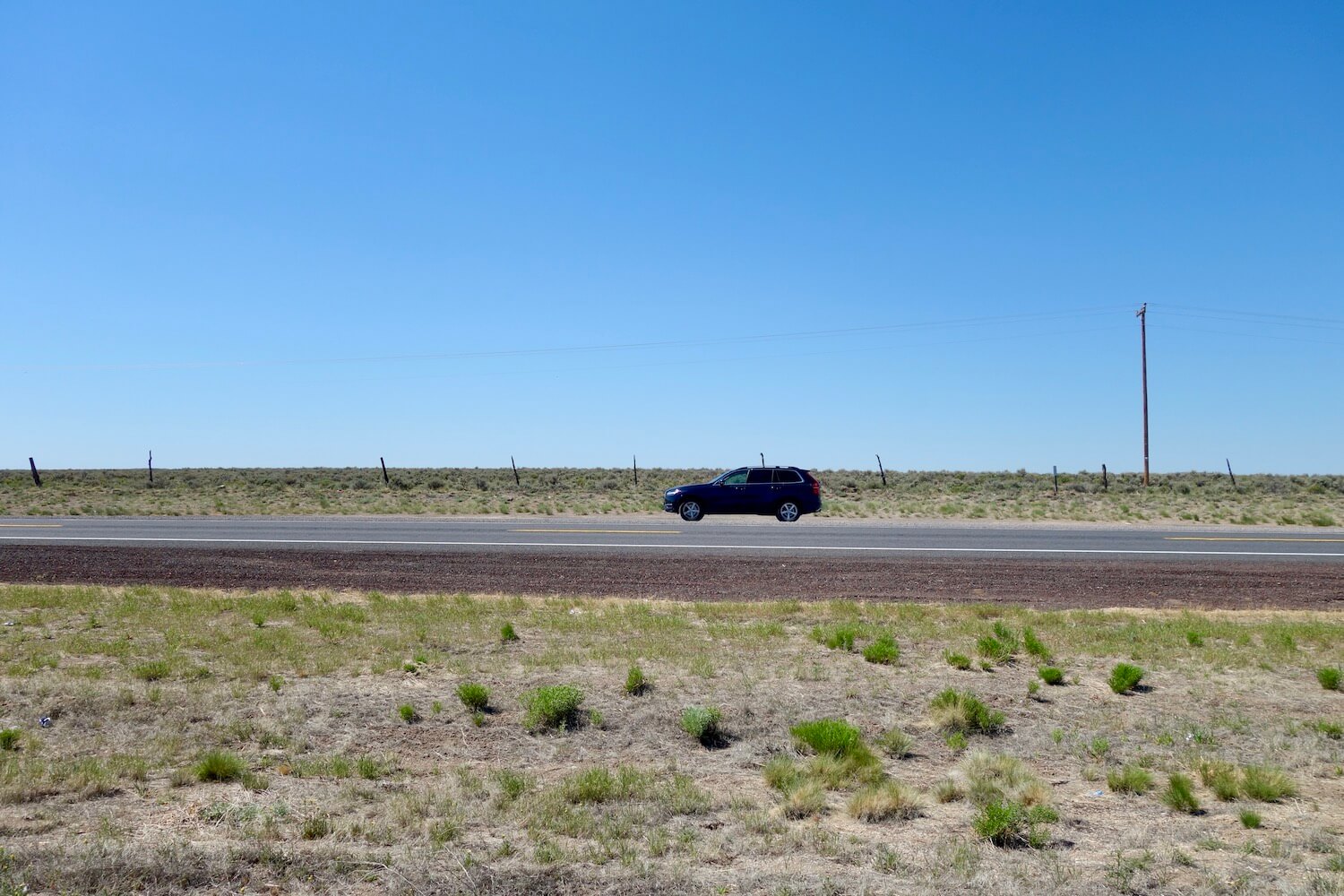 A lone blue SUV is parked on the side of the road amongst low brush and very dry looking landscape in Eastern Oregon.  There is a lone telephone pole amongst the blue sky.