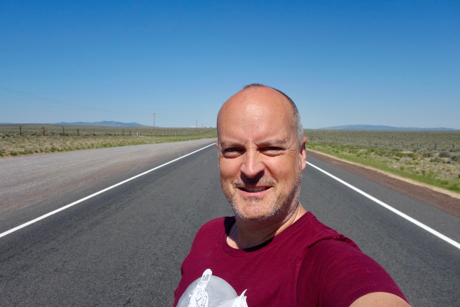 Matthew Kessi takes a selfie while on a road trip around Oregon.  He's standing in the middle of the highway and the white lines can be seen on both side.  He's wearing a red shirt and squinting a bit from the sunshine.  