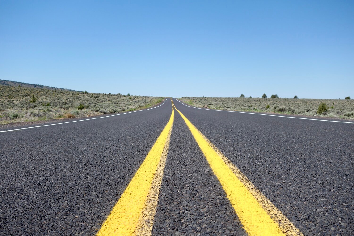 Two solid yellow lines flow through this photo leading away down a lonely blacktop highway in Eastern Oregon.  The sky is blue and the grassy terrain on either side is flat. 