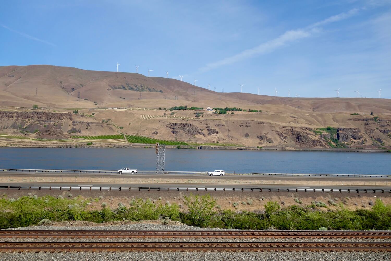 This shot of the Columbia River flowing through the Columbia River Gorge shows rusted train tacks, an interstate highway with two white cars driving and the mighty river.  The hills in the background are brownish colors under a blue sky. 