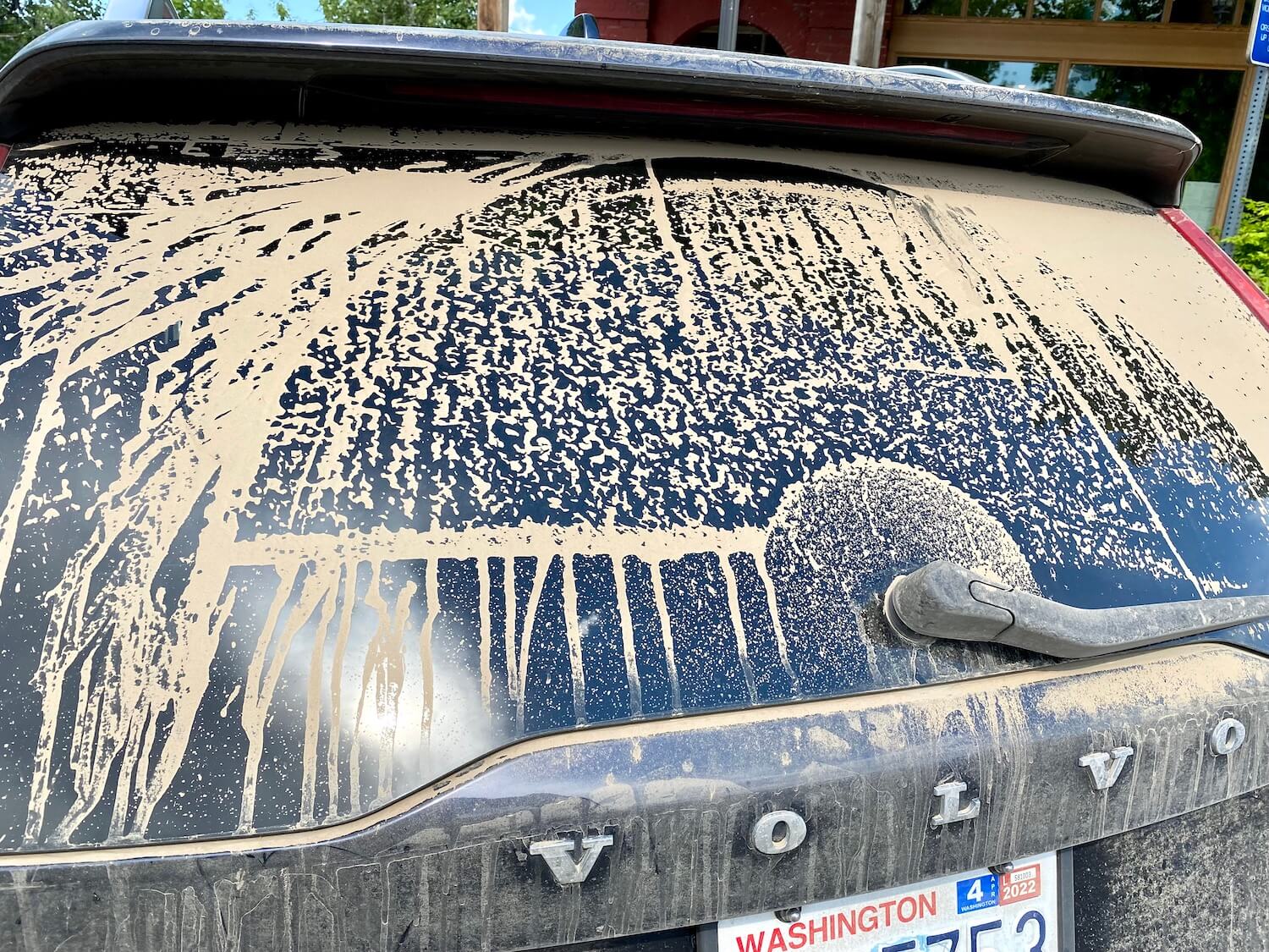 The back window of a Volvo is covered in mud and dust and the outline of the rear wiper seems to have made a trace through the brown goop.  A piece of a Washington license plate can be seen. 