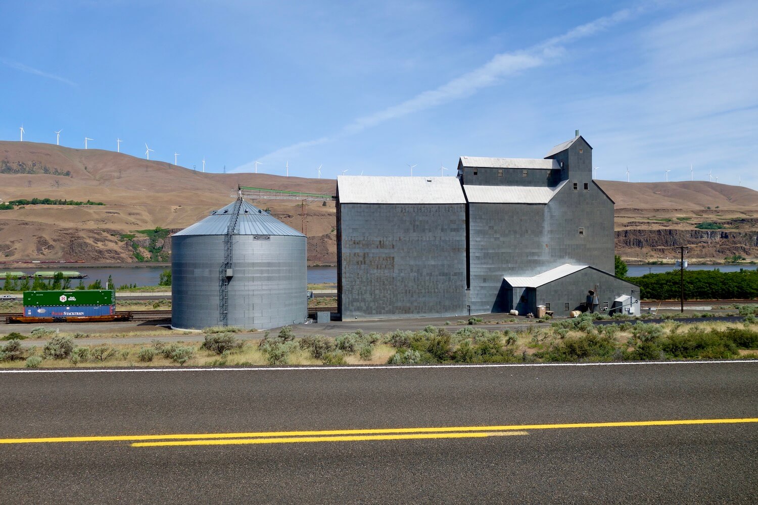 A large grain elevator and sheet metal covered barn rise up amongst a dry brownish hill.  The road has painted orange lines on blacktop that is smooth.  
