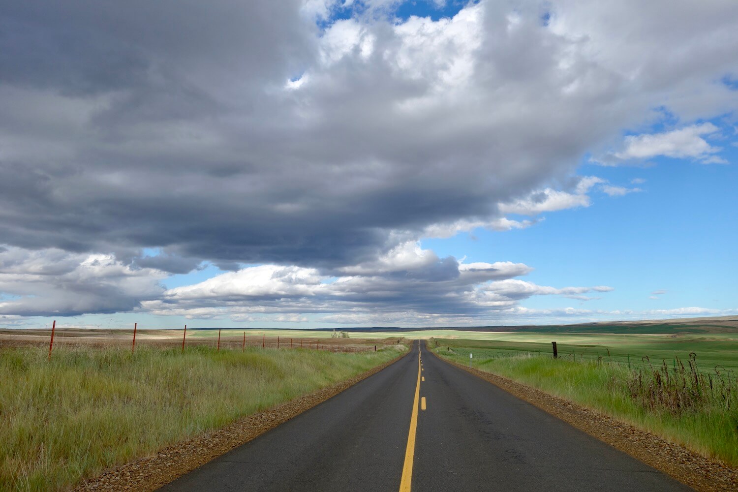 This shot taken on an Oregon road trip shows a lonely blacktop road with orange center lines underneath a dramatic system of clouds that look heavy with rain.  The fields on either side of the road are fenced in and full of green grass. 