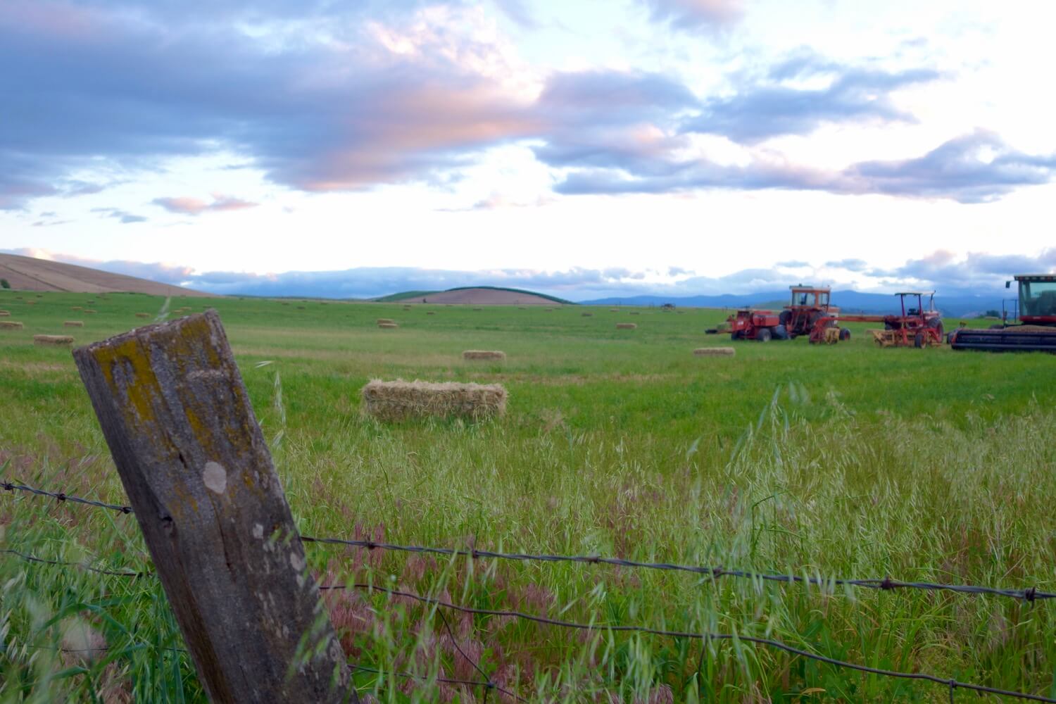This sunset scene taken from the side of a county road in near Dufur, Oregon focuses on a wooden fence post while a hay bale sits in a grassy field.  There are several pieces of farm machinery in the background. 