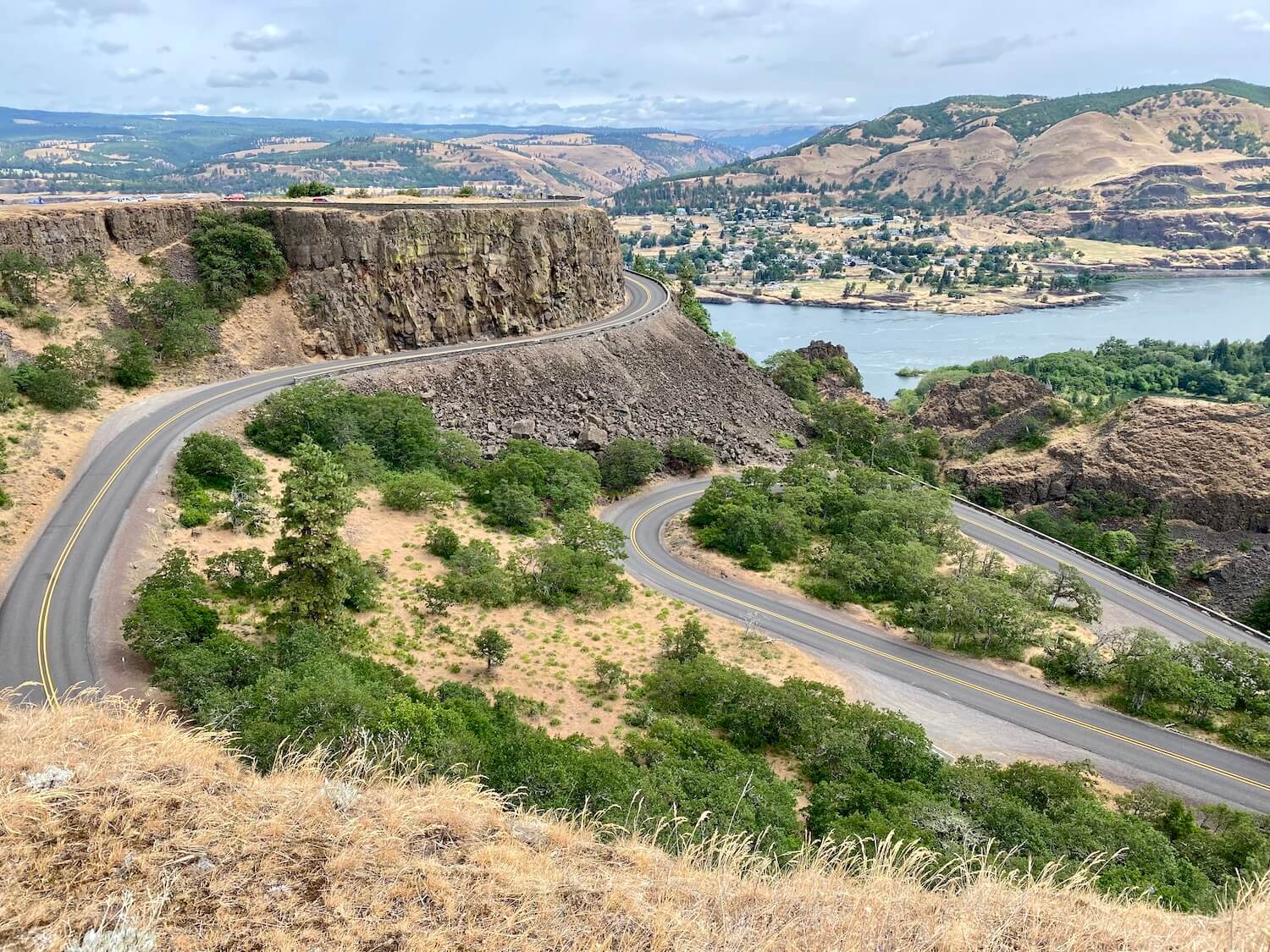 The curving road near the Rowena Lookout winds up a hill covered with yellow dried grasses and small green trees.  The Columbia River is visible in the background.  This photo was taken near The Dalles, Oregon. 