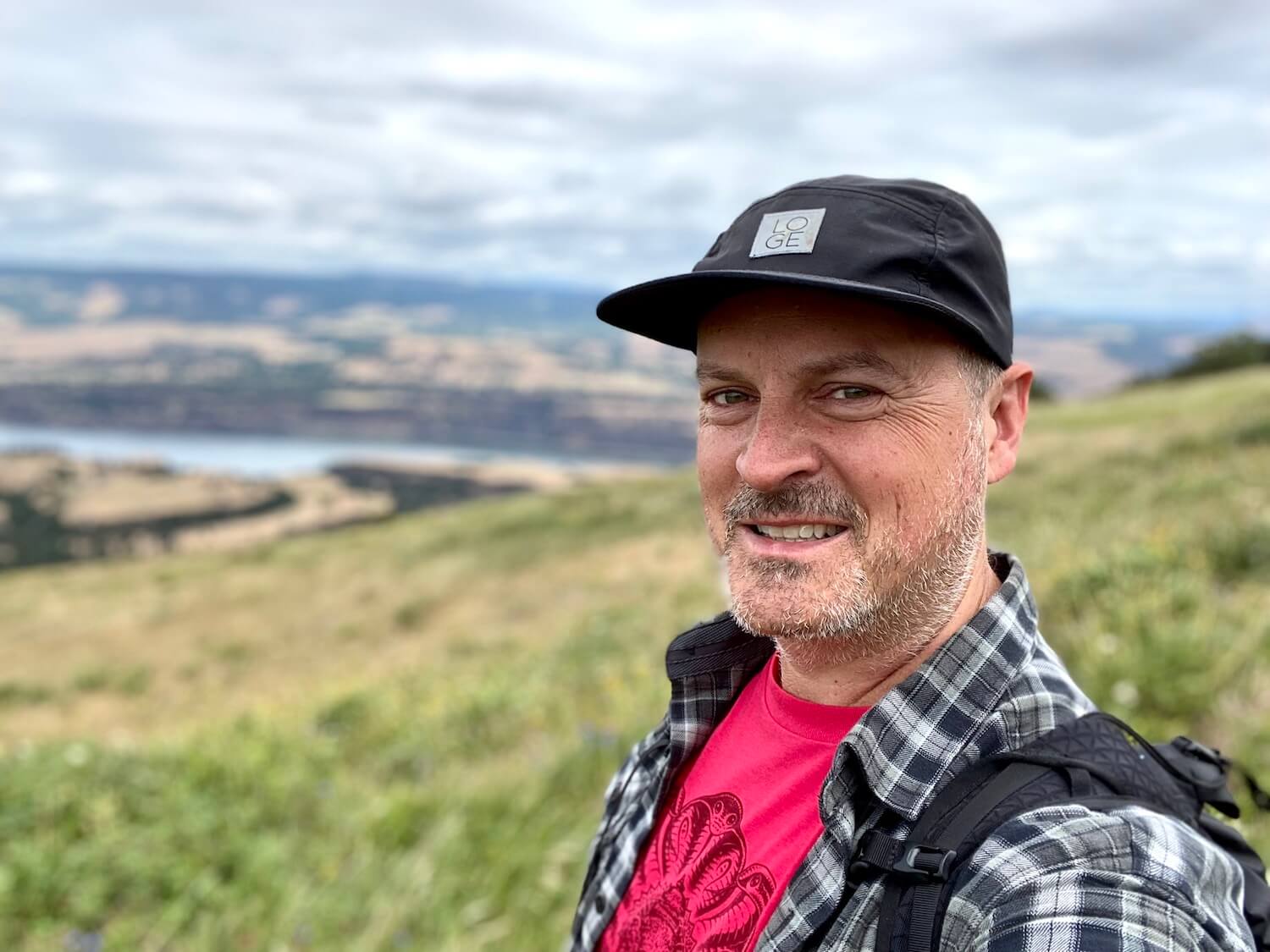 Matthew Kessi smiles for a selfie taken on his Oregon road trip.  He's wearing a bright red shirt covered with a black plaid shirt and a black cap on his head.  The background is out of focus but shows a green grassy meadow leading toward the Columbia River.