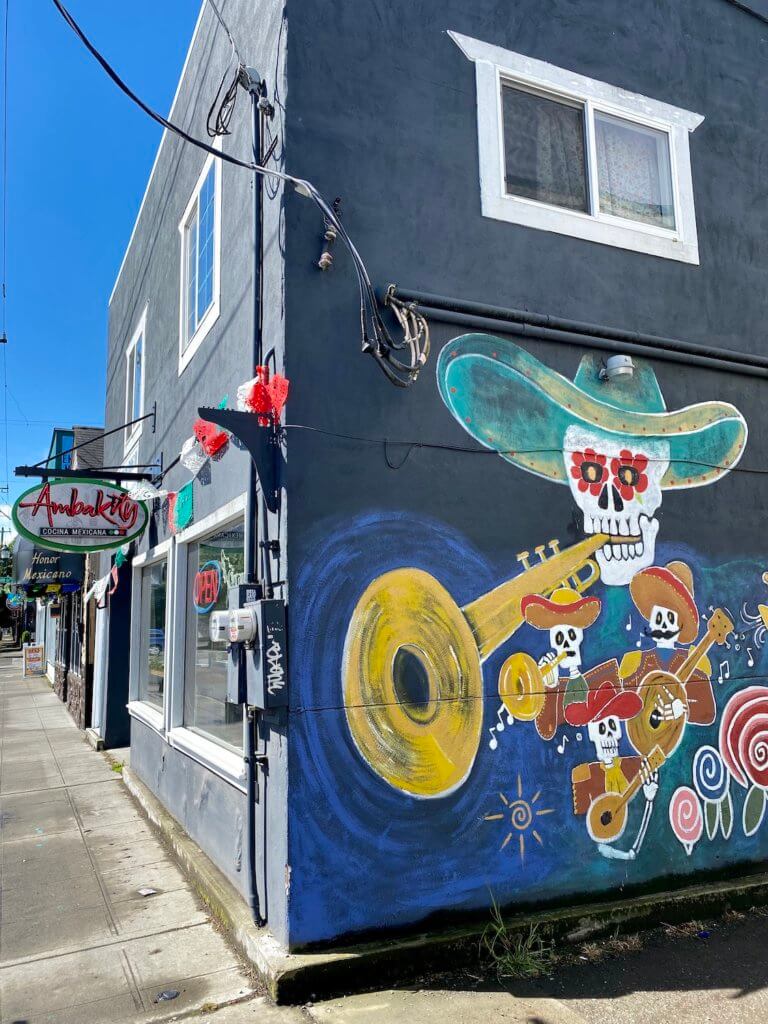 The side of a Mexican restaurant highlights a sugar skull wearing a sombrero playing a trumpet while the rest of the band, in smaller figures also play their instruments.  The rest of the building in the photo is the front of the restaurant, with a basic grey sidewalk and other signs in Spanish on the block in the South Park neighborhood of Seattle. 