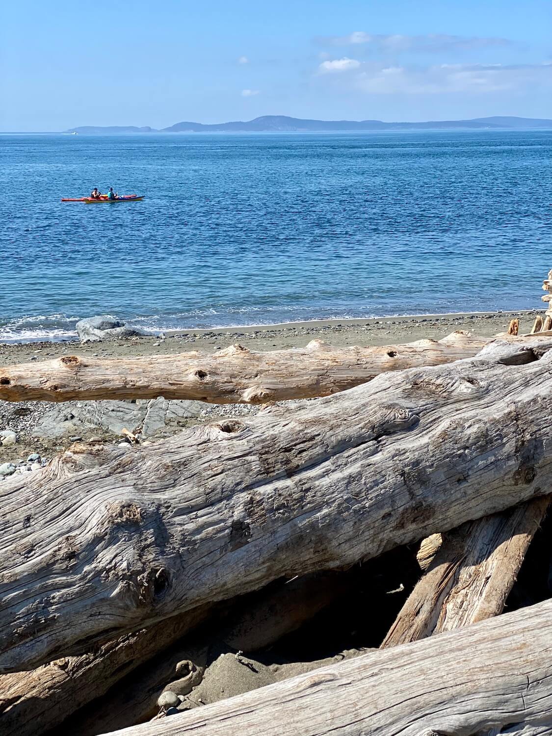 Two kayakers paddle in the waters off the coast of Deception Pass State Park. The beach is full of drift logs and lightly lapping waves while the background features the San Juan Islands under blue skies.