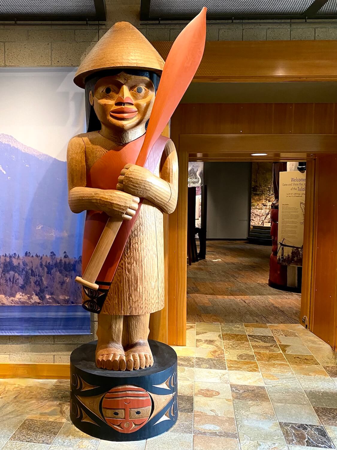 A tall carved wood figure stands to welcome visitors to the Hibulb Cultural Center.  The Indian is wearing a traditional cedar hat and holding a paddle to a canoe. 