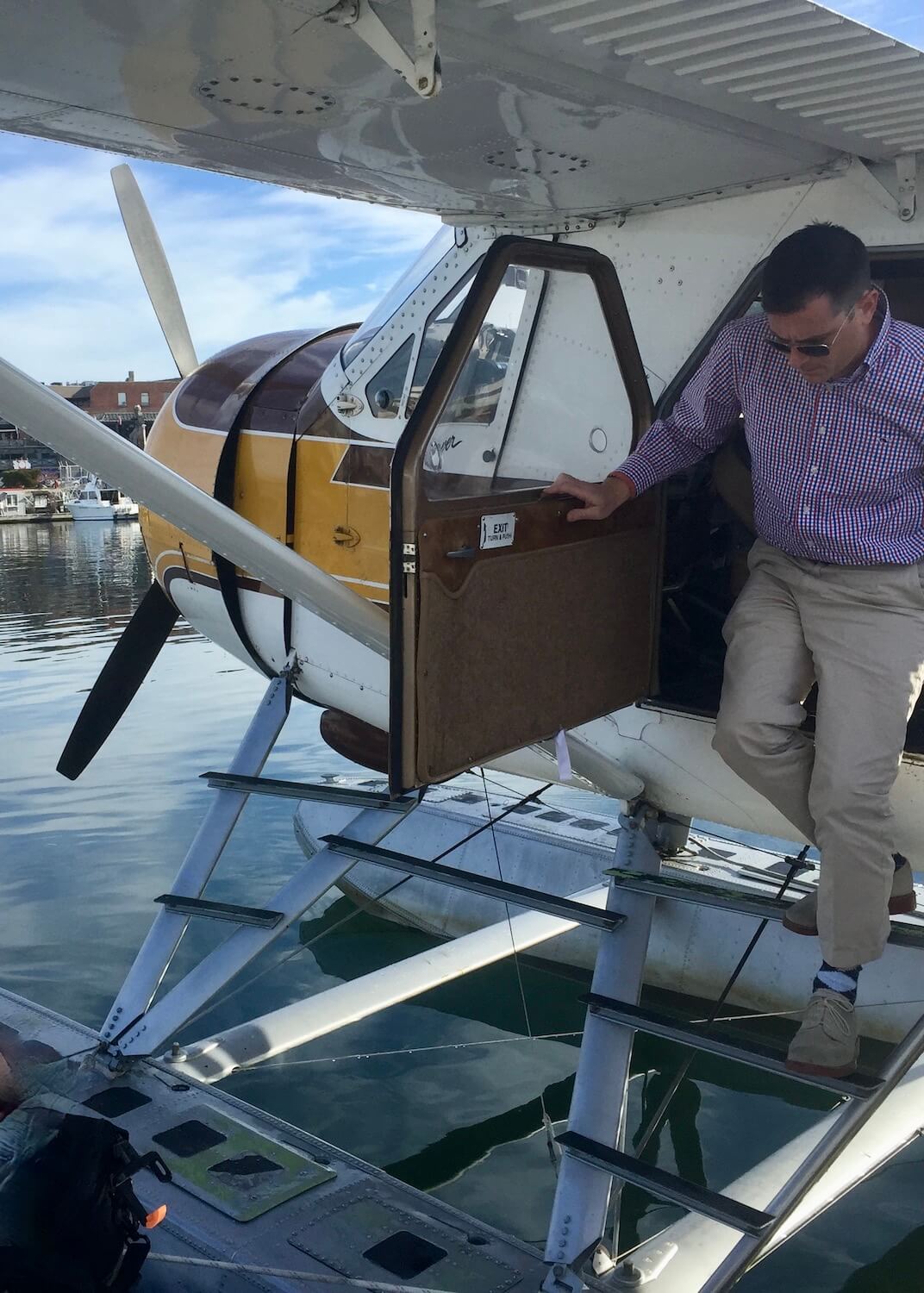 For those moving to Seattle a plane trip on Kenmore Air can be an exciting way to connect with nature.  Here a man with a purple checkered shirt steps out of a float plane with propellers in the front of the nose.