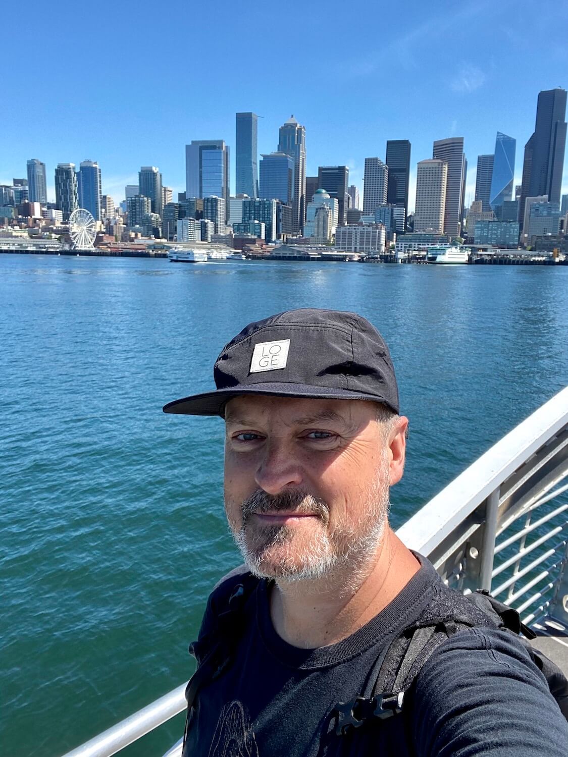 Matthew Kessi takes a selfie with the backdrop of Downtown Seattle behind him. He's on a boat with a metal railings and wearing a black t-shirt and hat.