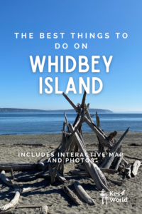 This Pinterest pin shows a pile of driftwood propped up to make a lean-to and shelter on the beach with blue water and distance land on the horizon in the background under blue sky.  The white writing shows things to do on Whidbey Island.