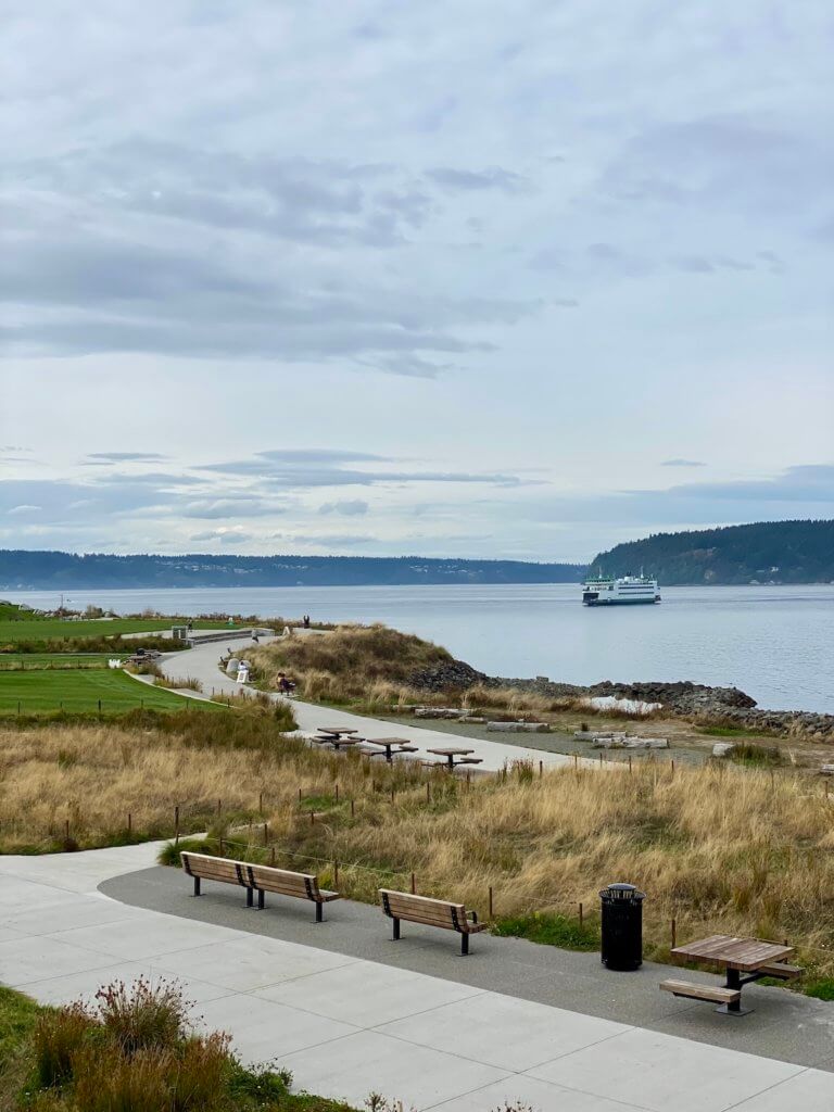 Dune Peninsula Park offers outstanding beauty to those wishing to have direct eyesight access to the Salish Sea. Here, a Washington State Ferry travels from Vashon Island to Tacoma Washington. A paved path winds through wavy seagrass.