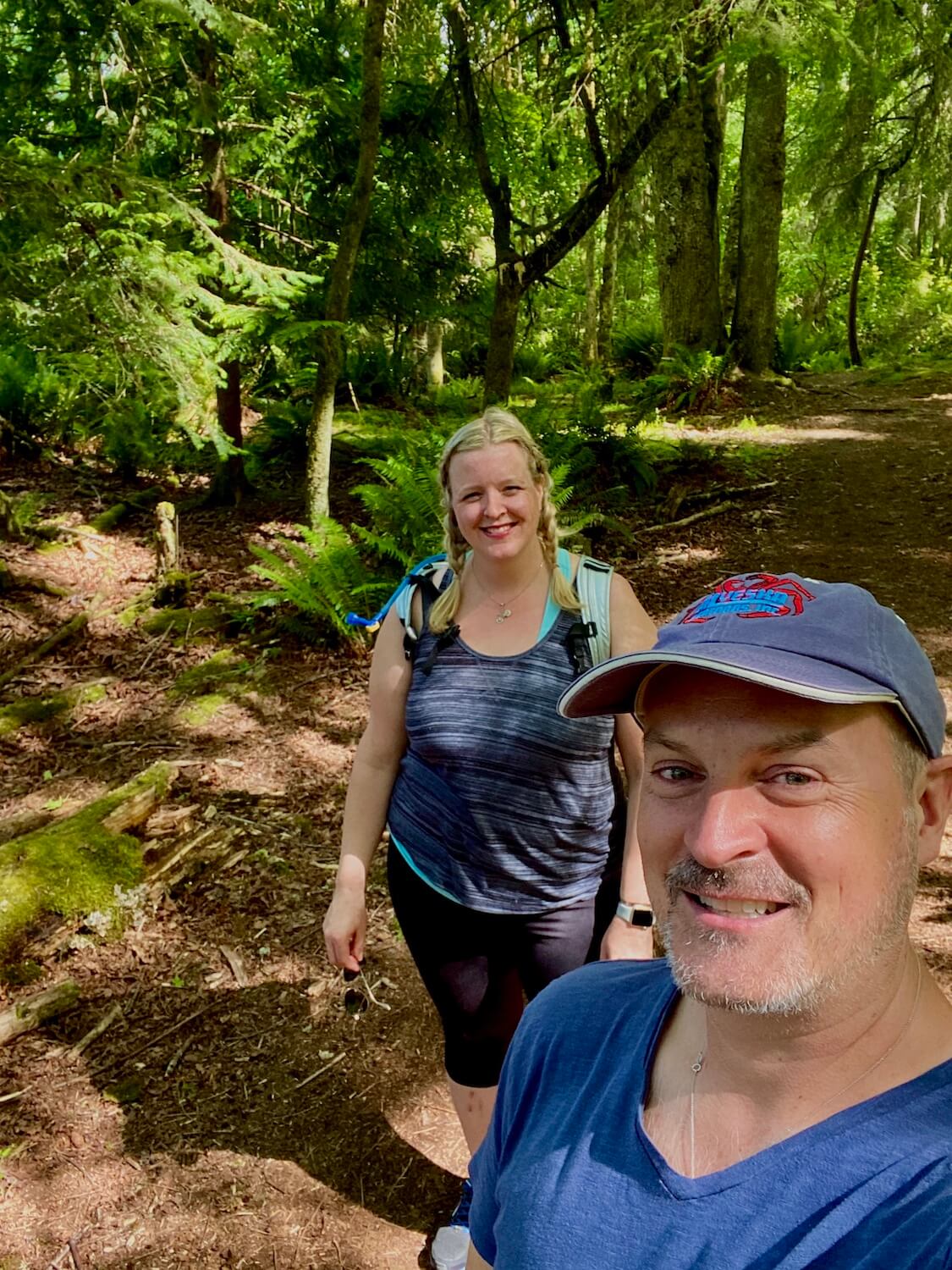 Matthew Kessi and Jeanne Jones are hiking in the trails deep in the forest of Point Defiance Park in Tacoma Washington.  Matthew wears a blue cap and t-shirt and is smiling while Jeanne is wearing a blue tank top.  They are surrounded by rich green trees with thick leaves and a sawdust trail on the forest floor. 