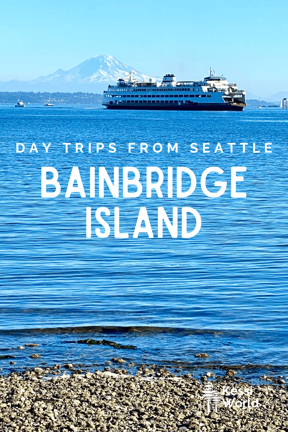 This Pinterest pin displays white letters that read "day trips from Seattle" and highlights Bainbridge Island. The photo shows a Washington State Ferry crossing below regal Mt. Rainier which rises up in the distance. The water has gentle ripples and a light wave brushes up onto the pebble beach.