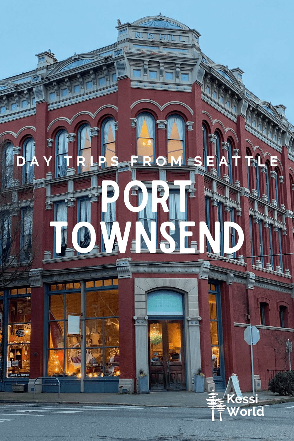 This Pinterest pin displays white letters that read "day trips from Seattle" and highlights Port Townsend. The photo shows an ornate Victorian-style three Storie building made from red bricks and cement decoration around the cornice of the buildings. The shop on the first floor has a orange glow from the candles lit in the window.