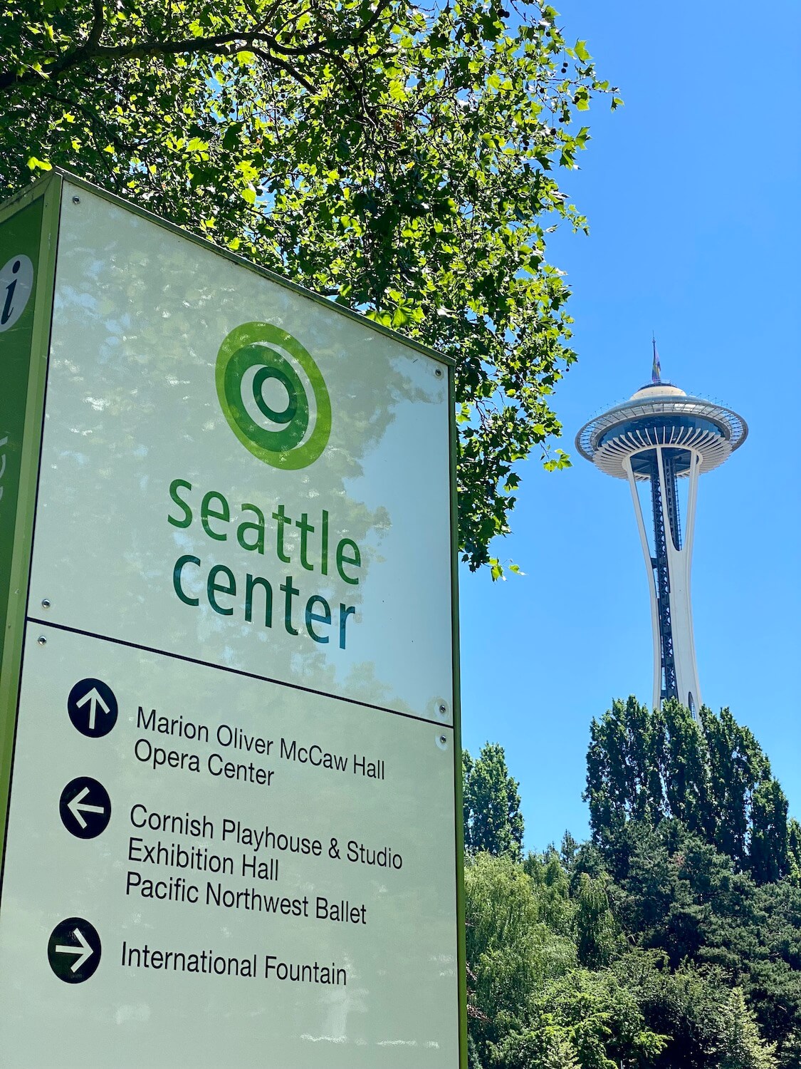Seattle Center is a fun outdoor thing to do in the Summer. This sign is green and white and says Seattle Center while the Space Needle rises in the background under bright blue skies.