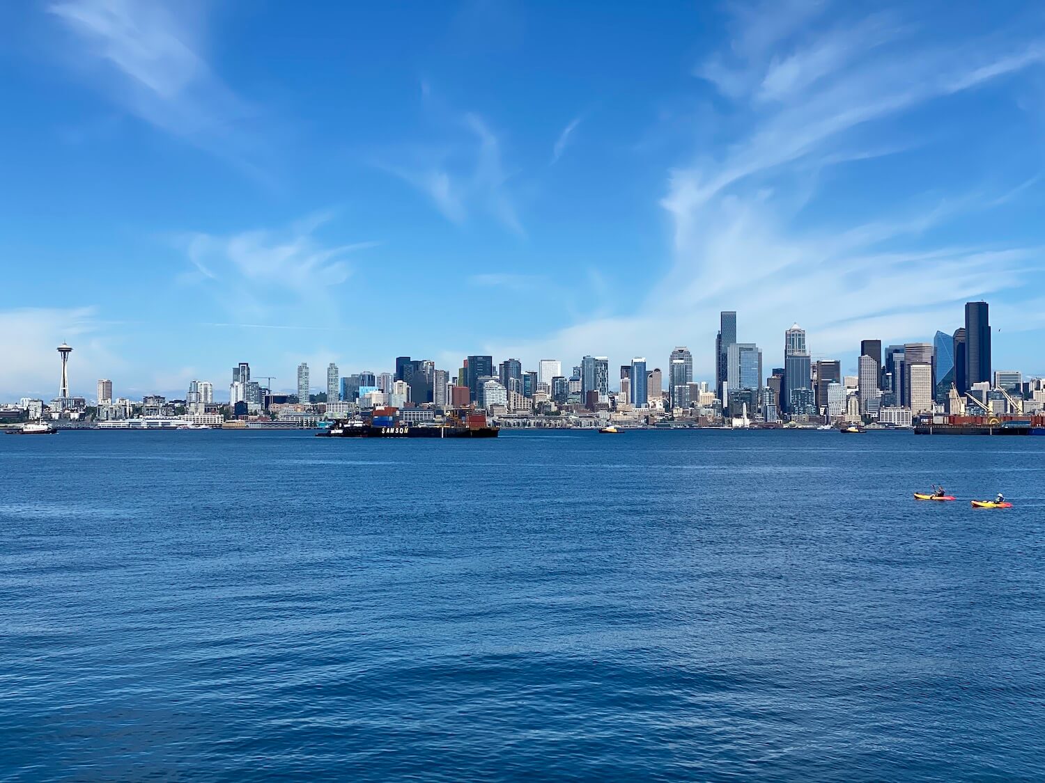 This panoramic view of the skyline of Seattle includes the Space Needle and the Columbia Center amongst other prominent buildings. The lush blue water of the Salish Sea is rolling with gentle waves while two kayakers in bright colored boats navigate the gentle waters.