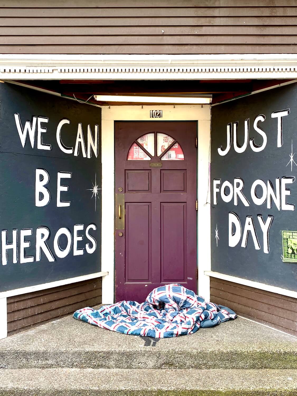 This shot shows a blanket on a doorstep of a building with street art that says, "We can be heroes just for one day."