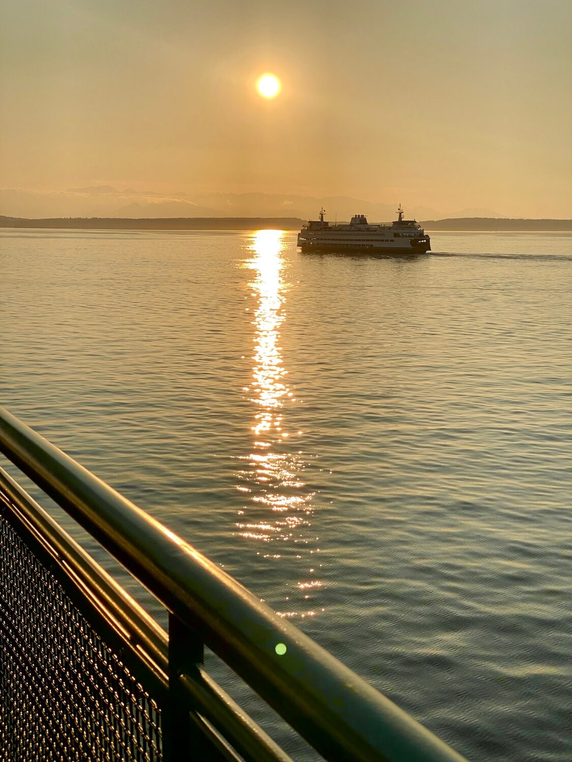 A Washington State Ferry crosses the Salish Sea, as seen from another ferry with a green railing.  The sun is setting over the Olympic Mountain range.