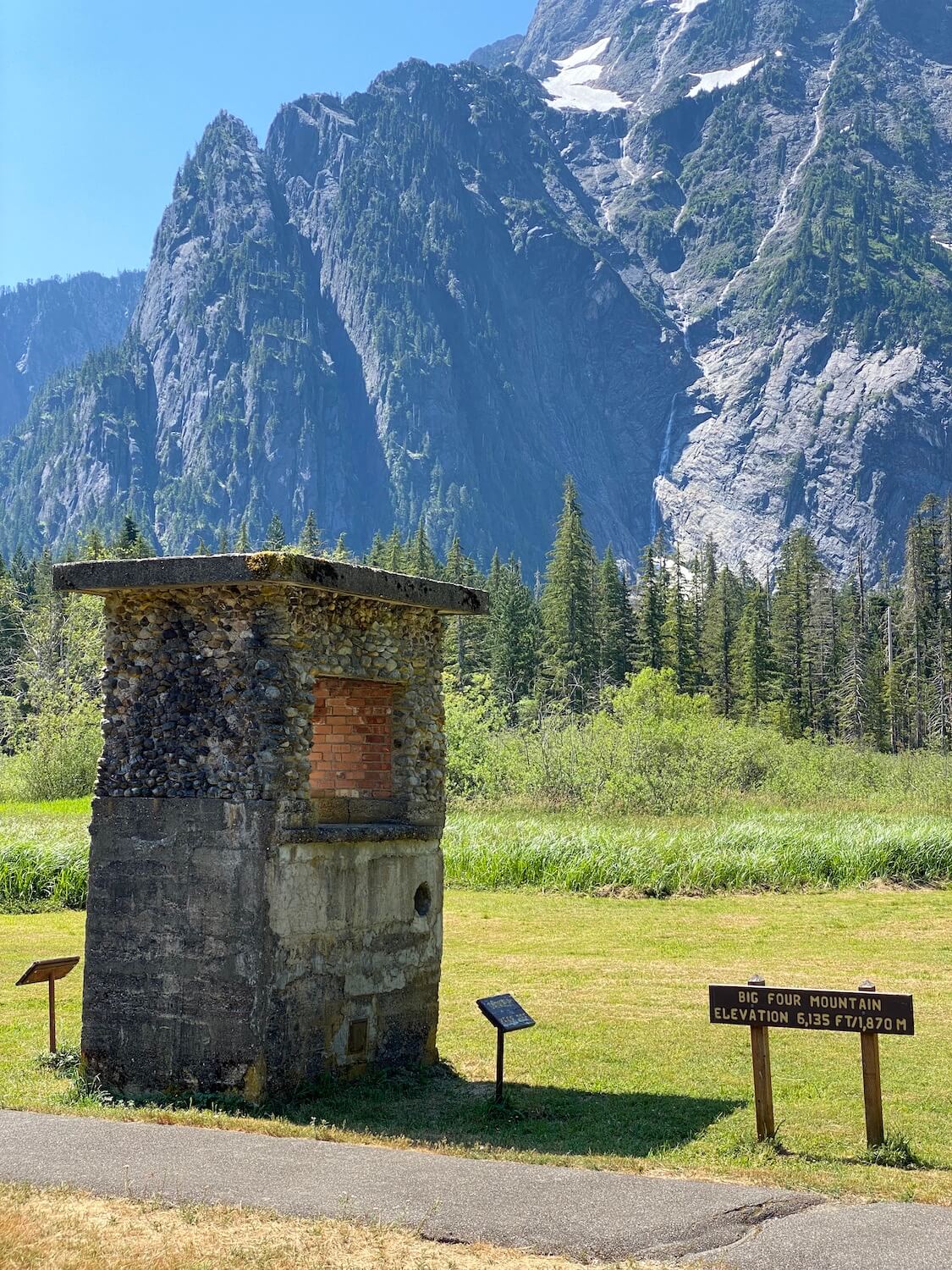 The remains of a historic lodge fireplace is highlighted next to a sign that marks Big Four Mountain. There is a grassy meadow that flows to a fir forest and then the towing mountain that still has patches of snow.